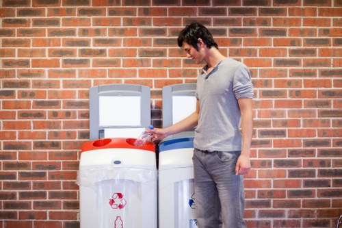 A young man with dark hair, wearing a light grey long-sleeved shirt and grey trousers, is standing outdoors beside two large white recycling bins placed on a paved area in front of a red brick wall. He is holding a clear plastic bottle and appears to be about to dispose of it into the bin on the left, which features a red recycling symbol on the front. The second bin, positioned to the right, is adjacent and also has a recycling symbol, indicating it is used for waste separation. The environment suggests an urban or residential setting typical for waste disposal and rubbish removal services. The image emphasizes proper recycling practices and waste management, aligned with services provided by companies such as Commercial Waste Waterloo serving the Waterloo area and its postal code, SE. The scene is well-lit, and the focus is on responsible waste disposal activities involving refuse segregation in the local community.