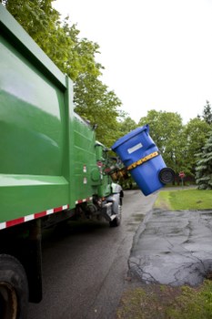 A green commercial waste collection truck positioned on a paved driveway or street, with its rear opening visible in the foreground. The truck is made of painted metal with a smooth finish and has reflective red and white striping along the lower side. A large blue wheelie bin is being lifted and emptied into the truck's hopper, with the bin's lid open and made of durable plastic. The surrounding environment includes a grassy area with mature trees in the background, and the scene appears to be on a residential or semi-urban street, possibly within the Waterloo area. The lighting is overcast, casting diffuse light that emphasizes the textures of the bin, metal surfaces, and paved ground. This image reflects commercial rubbish removal activity, likely managed by Commercial Waste Waterloo, illustrating the process of waste disposal or collection for local businesses or residents.