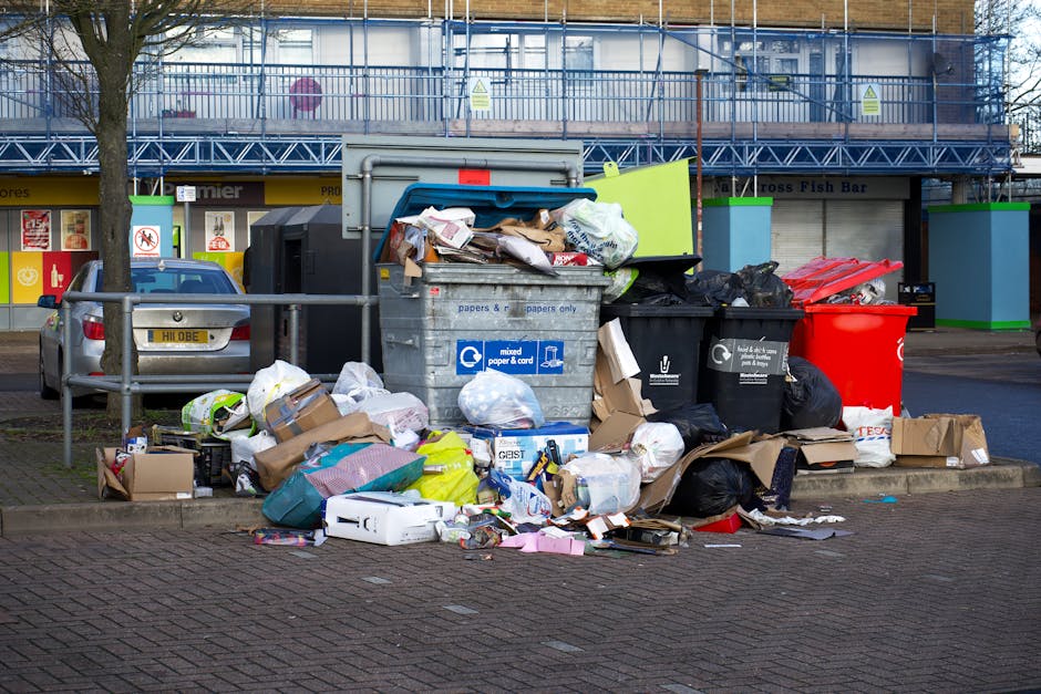A large pile of mixed waste and household rubbish, including cardboard boxes, plastic bags, paper, and packaging materials, is gathered around recycling bins on a paved urban sidewalk area. The bins a