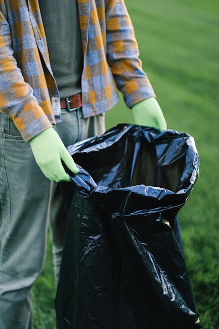 A person wearing a plaid shirt, grey pants, and lime green gloves is holding open a large black garbage bag lined with a shiny black plastic interior, preparing it for waste collection. The individual