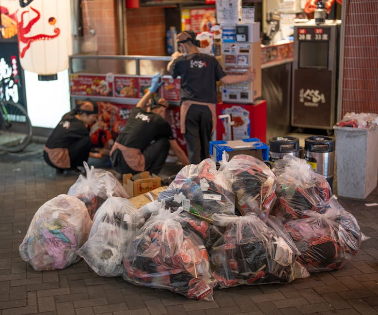 An alleyway cluttered with accumulated waste materials, featuring large black and white plastic bags filled with rubbish in the foreground, with some spills on the pavement beneath. Behind the bags, t
